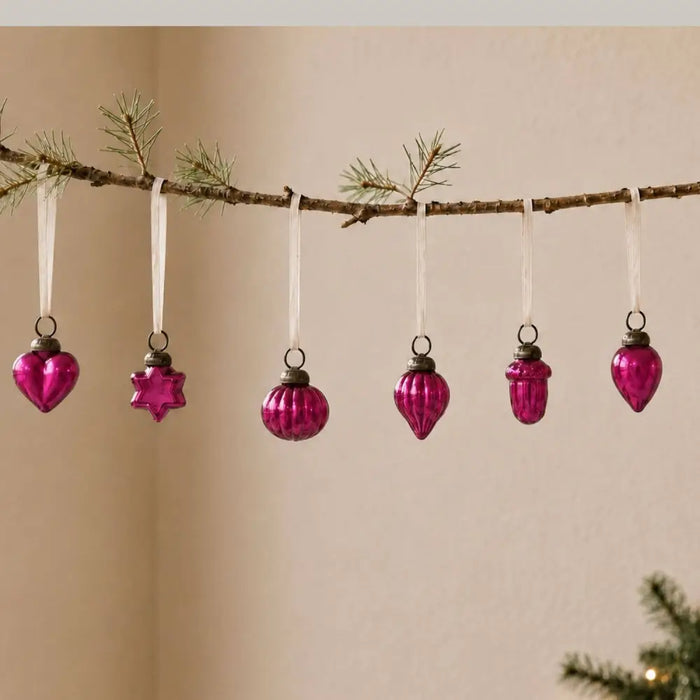 Small fuchsia mouth-blown glass baubles with metallic caps hang from white ribbons on a pine branch