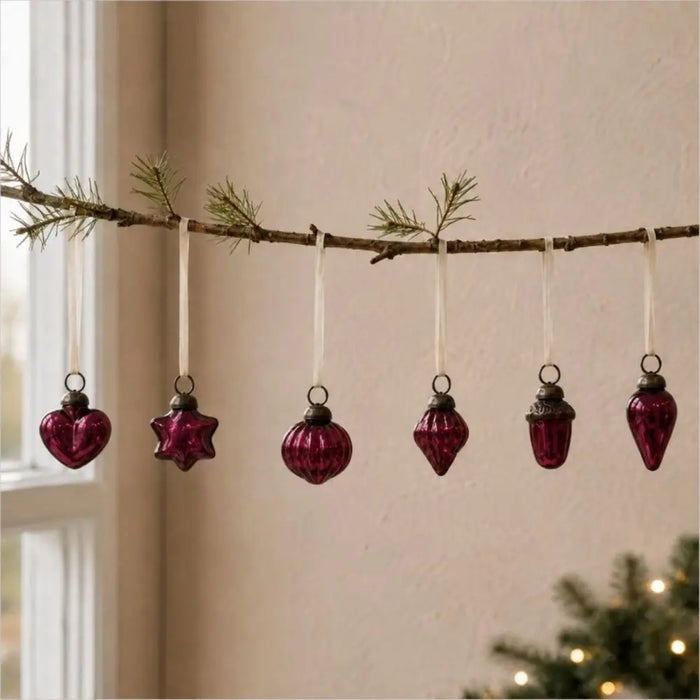 A set of six deep purple mouth-blown glass baubles with metallic caps hanging from white ribbons on a rustic branch