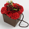 A soap flower bouquet with red roses and carnations green leaves and small yellow flowers in a brown paper box