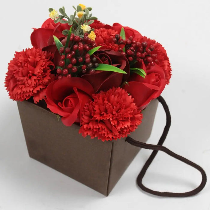 A soap flower bouquet with red roses and carnations green leaves and small yellow flowers in a brown paper box