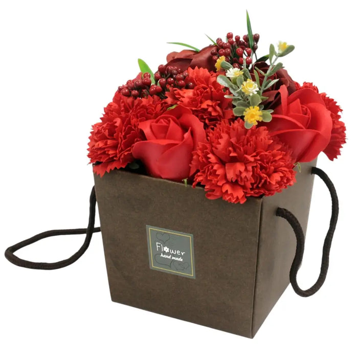 A soap flowers bouquet with red roses and carnations in a brown paper gift box with black handles