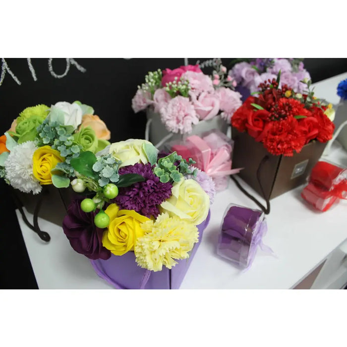 A soap flower bouquet with yellow roses white carnations and purple chrysanthemums in a purple box with green leaves