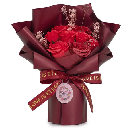 A soap flower bouquet of red roses wrapped in maroon paper with a matching ribbon and a gift card tag