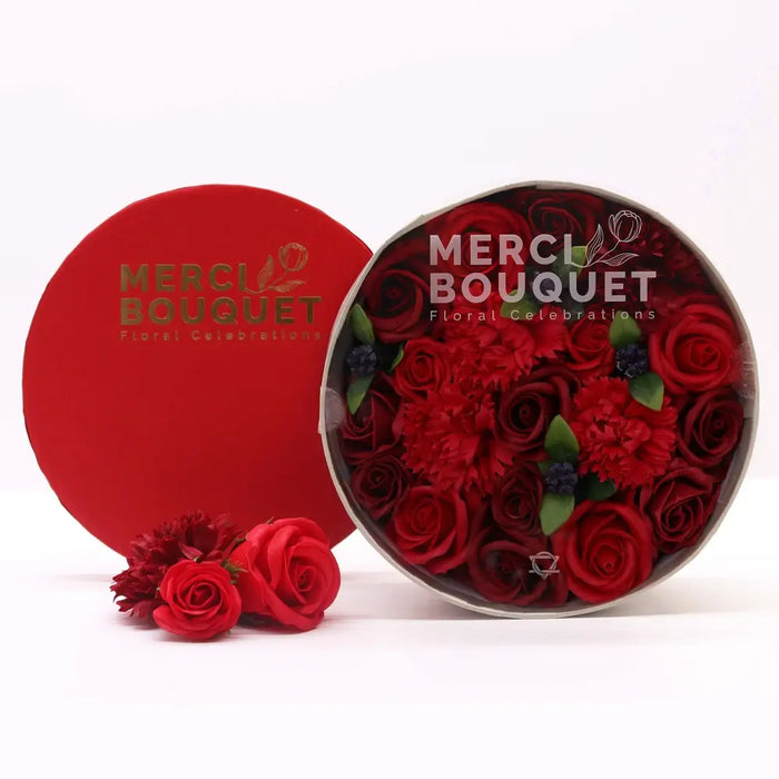 A vibrant red circular soap flower gift box filled with artificial red roses and carnations by merci bouquet floral celebrations