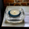 Clear glass tealight holder with a teal inner ring holding a lit white tea light candle in a square design