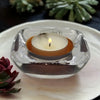 A clear glass tealight holder with a square curved design holding an orange rimmed glass tealight candle