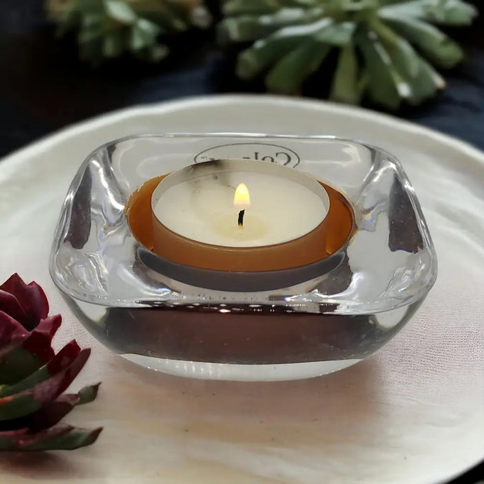 A clear glass tealight holder with a square curved design holding an orange rimmed glass tealight candle