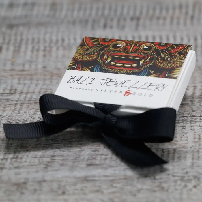 A white jewelry box with a balinese mask design in reds and golds tied with black ribbon for sterling silver and 18ct yellow gold earrings