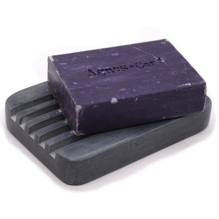 A rectangular purple soap bar with agnes court rests on a grey soapstone soap dish with deep ridges and slats