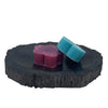 Two colorful flower shaped soap bars in deep purple and teal on a dark petrified wood soap dish slice