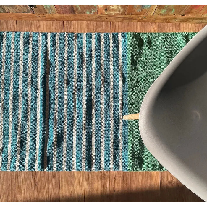A handwoven indian cotton rug with vertical teal white and green stripes in a cozy indoor setting