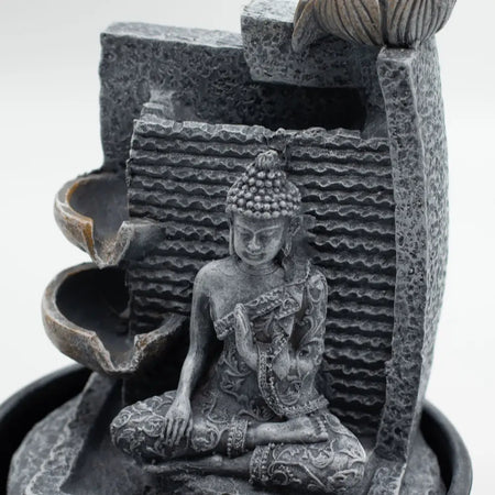 Gray stone buddha figurine with intricate carvings seated in meditation in a lotus flower cascading tabletop water feature