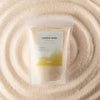 The Window, Milford Haven Blush Bloom fragranced candle sand in a 300g pouch, surrounded by golden wax granules.