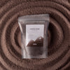 The Window, Milford Haven Millionaire Gold chocolate brown candle sand in a pouch set against a textured sand spiral.