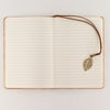 The Window, Milford Haven A5 notebook open to lined pages with a brass-coloured leaf bookmark on a cork effect cover.