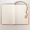 The Window, Milford Haven A5 notebook open to lined paper pages with a brass leaf bookmark on a red leather cover.