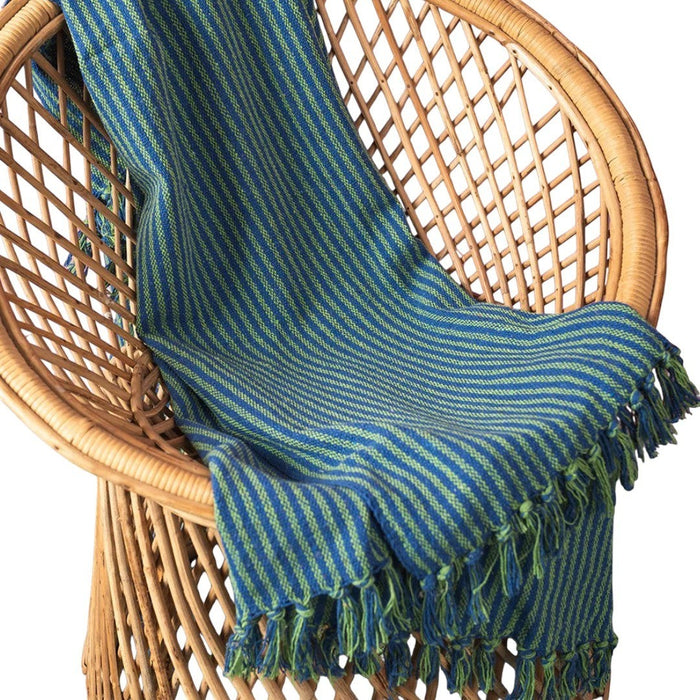 The Window, Milford Haven green and blue striped recycled cotton throw with tasselled fringe draped on a wicker chair.