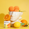 The Window, Milford Haven Vitamin C bath bomb in a white face-shaped bowl with fresh oranges and a lemon on yellow