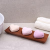Three heart-shaped bath bombs in pink and lavender resting on a naseberry wood soap dish tray