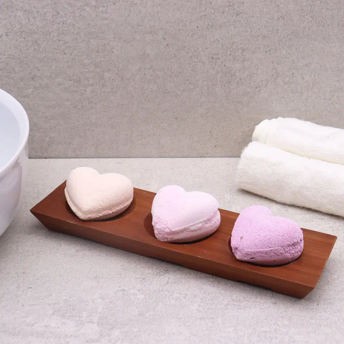 Three heart-shaped bath bombs in pink and lavender resting on a naseberry wood soap dish tray