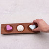 A wooden naseberry wood tray with three circular indentations holding colorful heart and flower shaped soap bars