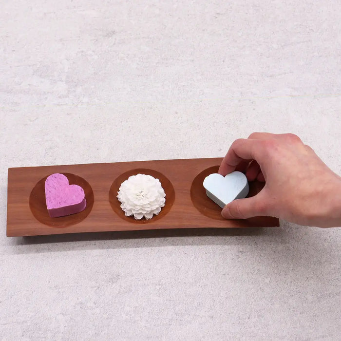 A wooden naseberry wood tray with three circular indentations holding colorful heart and flower shaped soap bars
