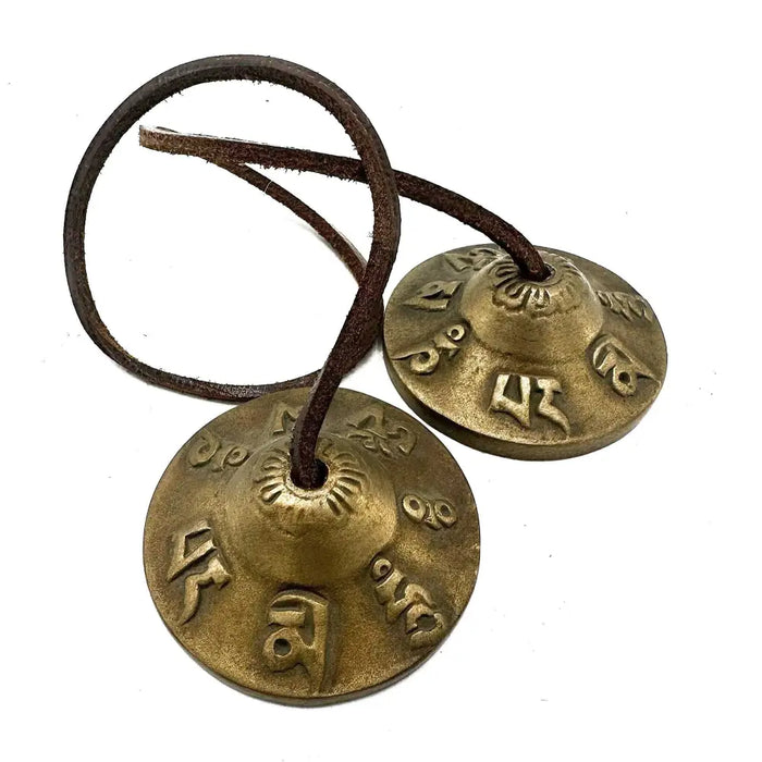 Brass-colored tibetan tingsha prayer bells with engraved sanskrit script hanging from dark leather cord lucky symbols