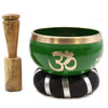 Green tibetan singing bowl set with gold om symbol and brass rim on black silver cushion next to wooden mallet