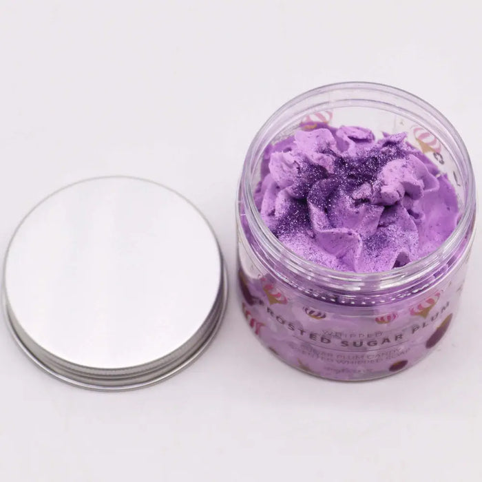 A clear glass jar with silver lid holds purple glittery whipped soap bath scrub labeled roasted sugar plum frosted sugar plum