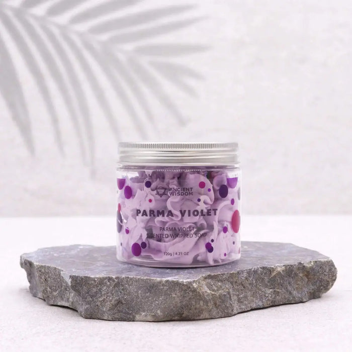 A clear glass jar with silver lid holds lavender whipped soap speckled with purple and pink dots labeled parma violet for skin feeling