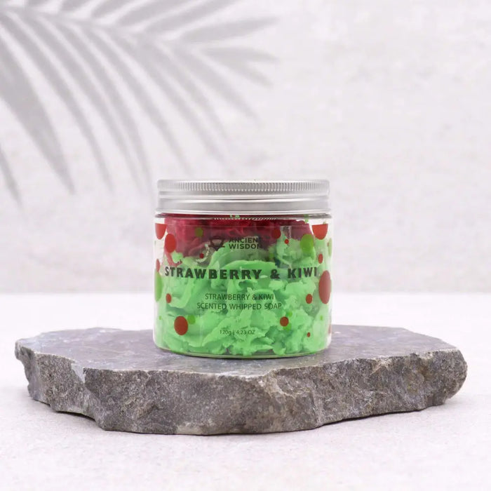 A clear glass jar with a silver lid holds green strawberry and kiwi scented whipped soap with red fruit pieces