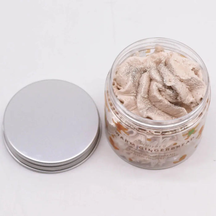A clear glass jar with silver lid holding light beige whipped soap flecked with glitter in warm gingerbread scent
