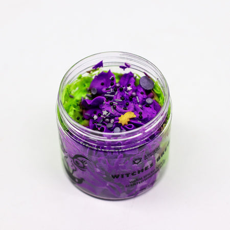 A clear glass jar of witches brew whipped soap bath bombs in purple and green with black and silver glitter decorations