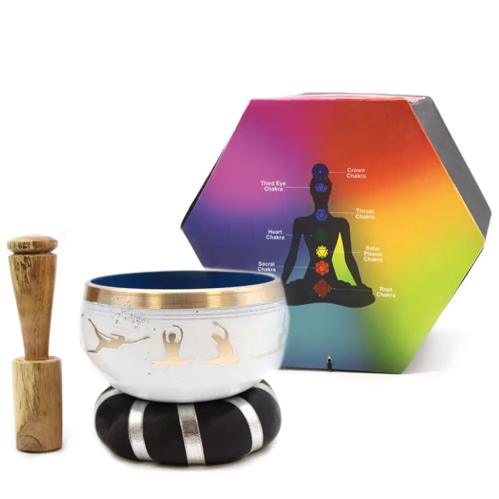 White singing bowl with gold rim and engraved yoga moves figures on a black and silver cushion beside wooden mallet