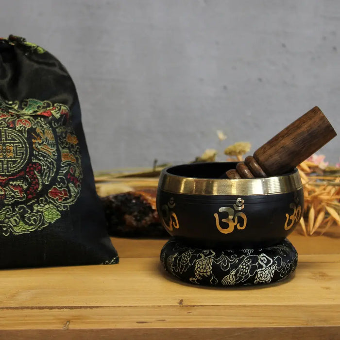 Black tibetan singing bowl set with gold om symbols, brass rim resting on an ornate black cushion in the singing bowl set